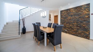 A modern dining area with a wooden table and black chairs, a staircase with a glass railing, and a stone wall. - Casa no Sol | 3 Bedrooms | Modern | Almancil (Almancil)