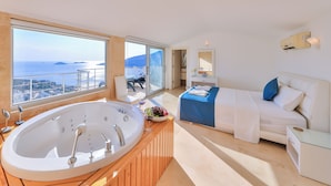 Quarto de hotel moderno com uma banheira grande, cama com colcha azul e branca e vista para o mar e ilhas distantes.