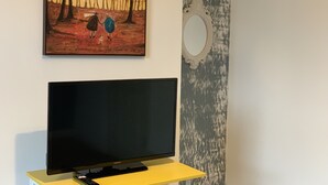 Un televisor sobre un soporte amarillo con un control remoto, un cuadro de dos personas caminando en un bosque y un espejo en la pared.