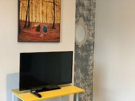 Uma televisão sobre um suporte amarelo, um controle remoto, uma pintura de duas pessoas caminhando em uma floresta e um espelho na parede.