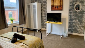 Un dormitorio con cama, armario, televisor sobre un soporte y un cuadro en la pared.