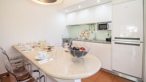 Een keuken met een witte koelkast, een magnetron, een spoelbak en een mand met fruit op het aanrecht.