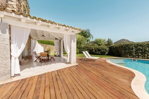 Une terrasse en bois avec une piscine, un patio couvert comprenant une table et des chaises, et une vue sur le paysage environnant.