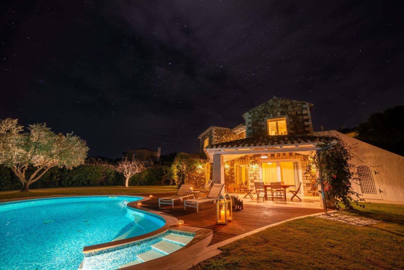 Vista noturna de uma casa com piscina, área de estar externa e fachada de pedra.