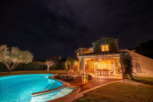A nighttime view of a house with a swimming pool, outdoor seating, and a stone facade.