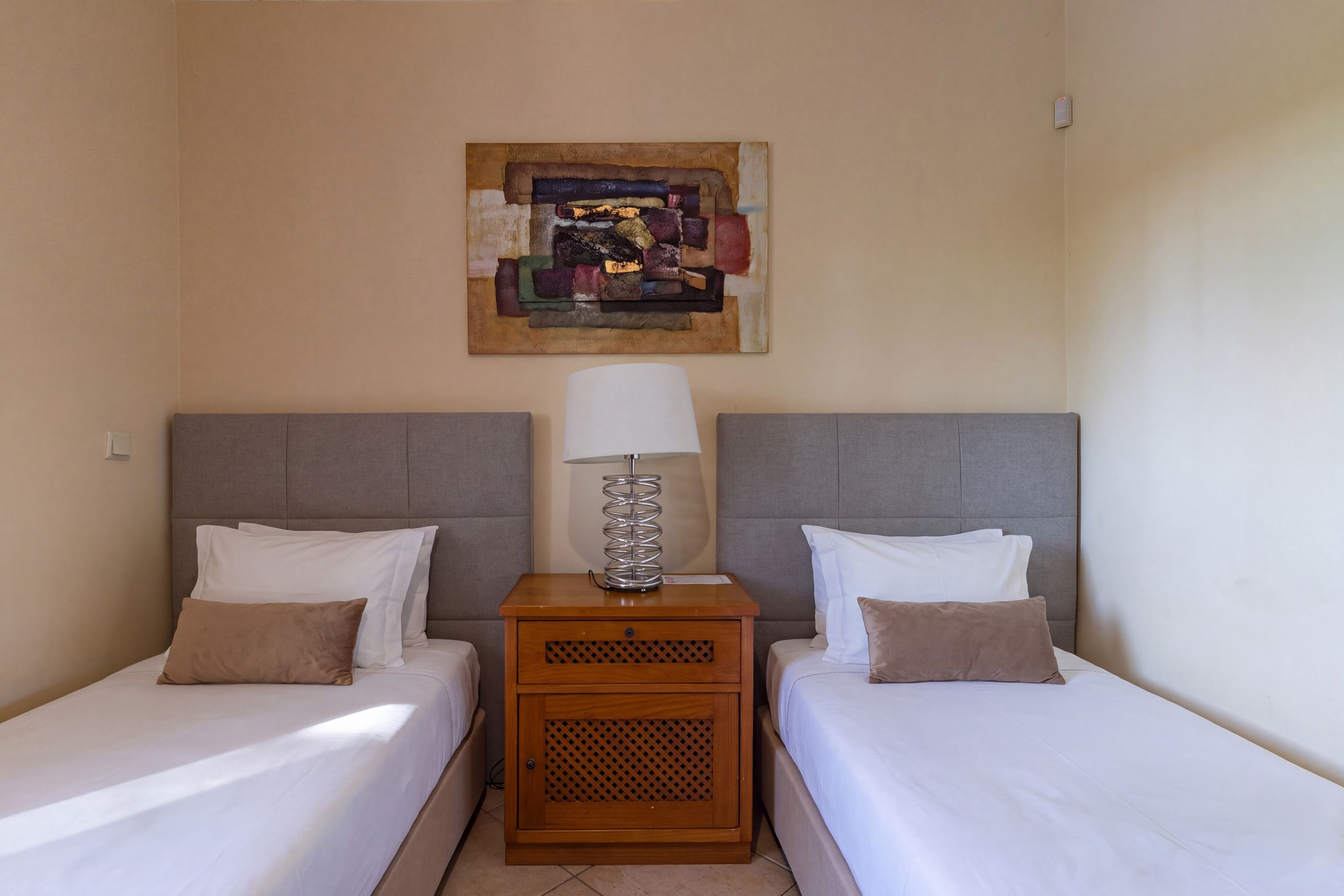 A hotel room with two single beds, a wooden nightstand with a lamp, and a wall-mounted artwork.