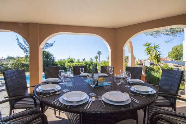 A round dining table with chairs, set for a meal, surrounded by arches with a pool and greenery in the background.