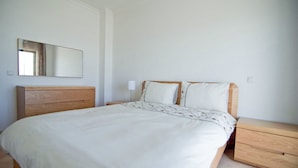 Een slaapkamer met een houten bed, wit beddengoed, een nachtkastje, een spiegel en een lichtschakelaar op de muur.