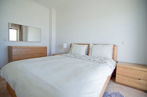 A bedroom with a wooden bed, white bedding, a nightstand, a mirror, and a light switch on the wall.