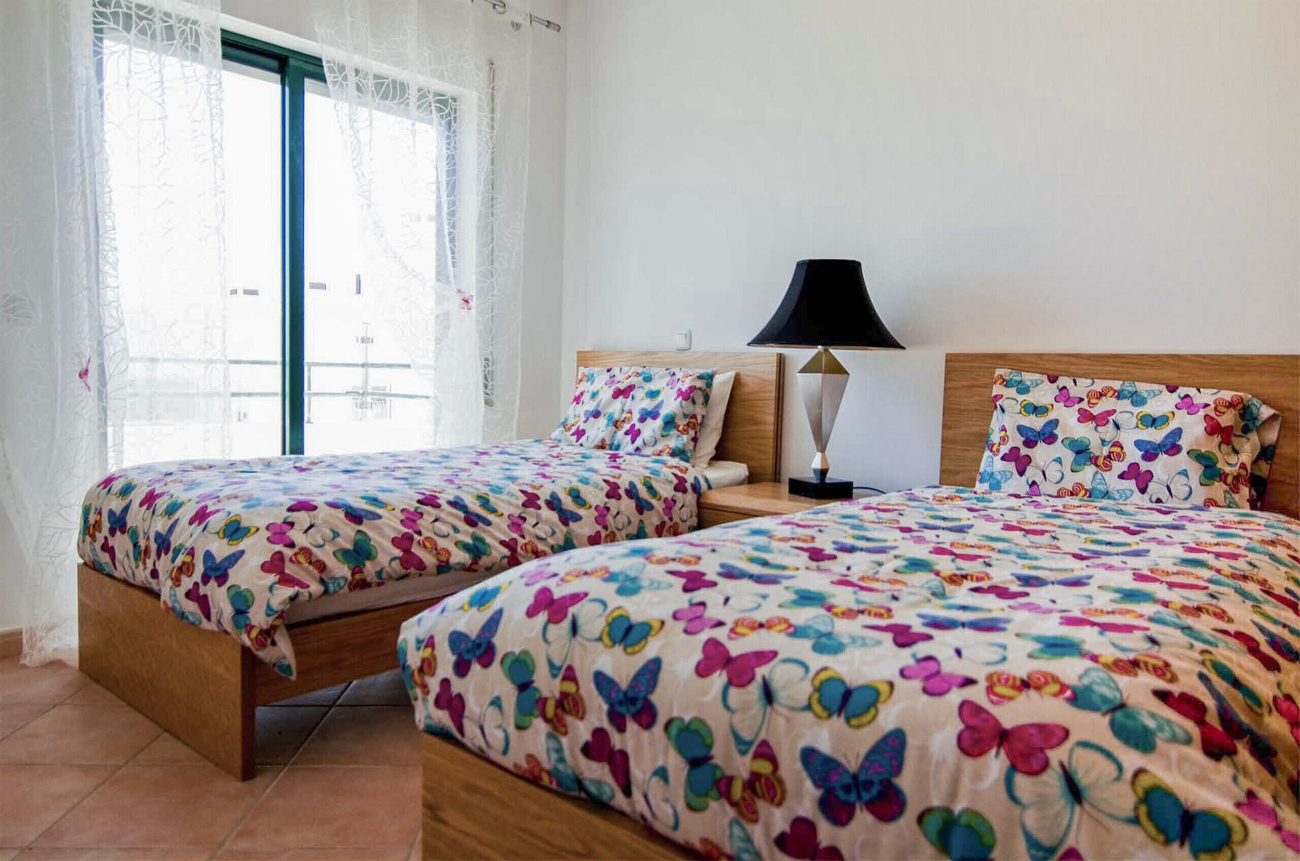 A room with two beds, a nightstand with a lamp, and a window with a curtain.