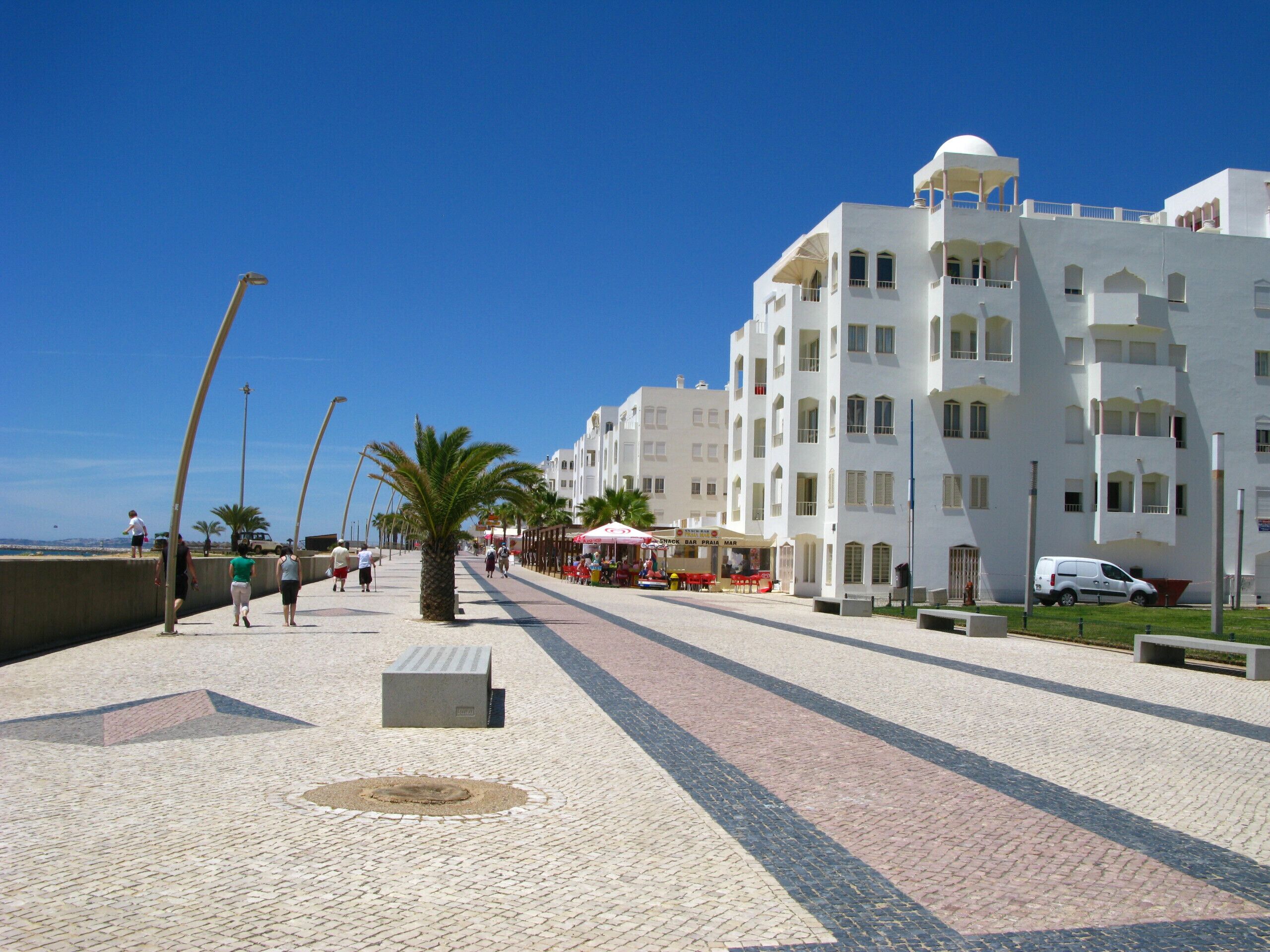A coastal promenade with white buildings, palm trees, and outdoor seating areas.
