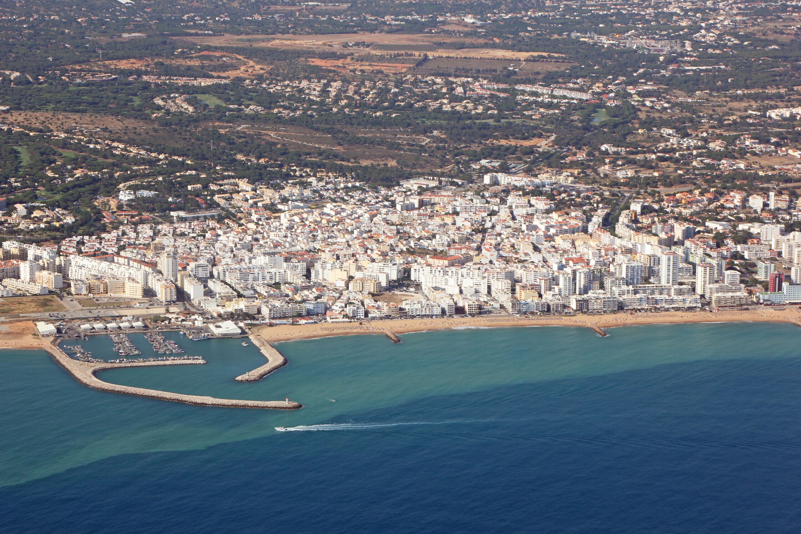 An aerial view of a coastal city with dense urban development, a marina, and a curved breakwater.