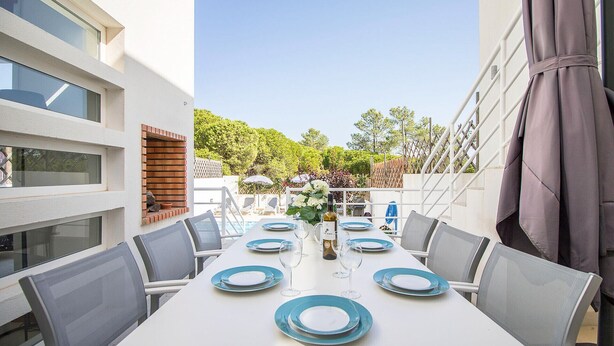 Un coin repas avec une table blanche dressée pour six personnes, des chaises d’extérieur et une vue sur un jardin et des arbres.
