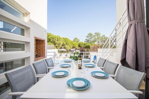 Un coin repas avec une table blanche dressée pour six personnes, des chaises d’extérieur et une vue sur un jardin et des arbres.