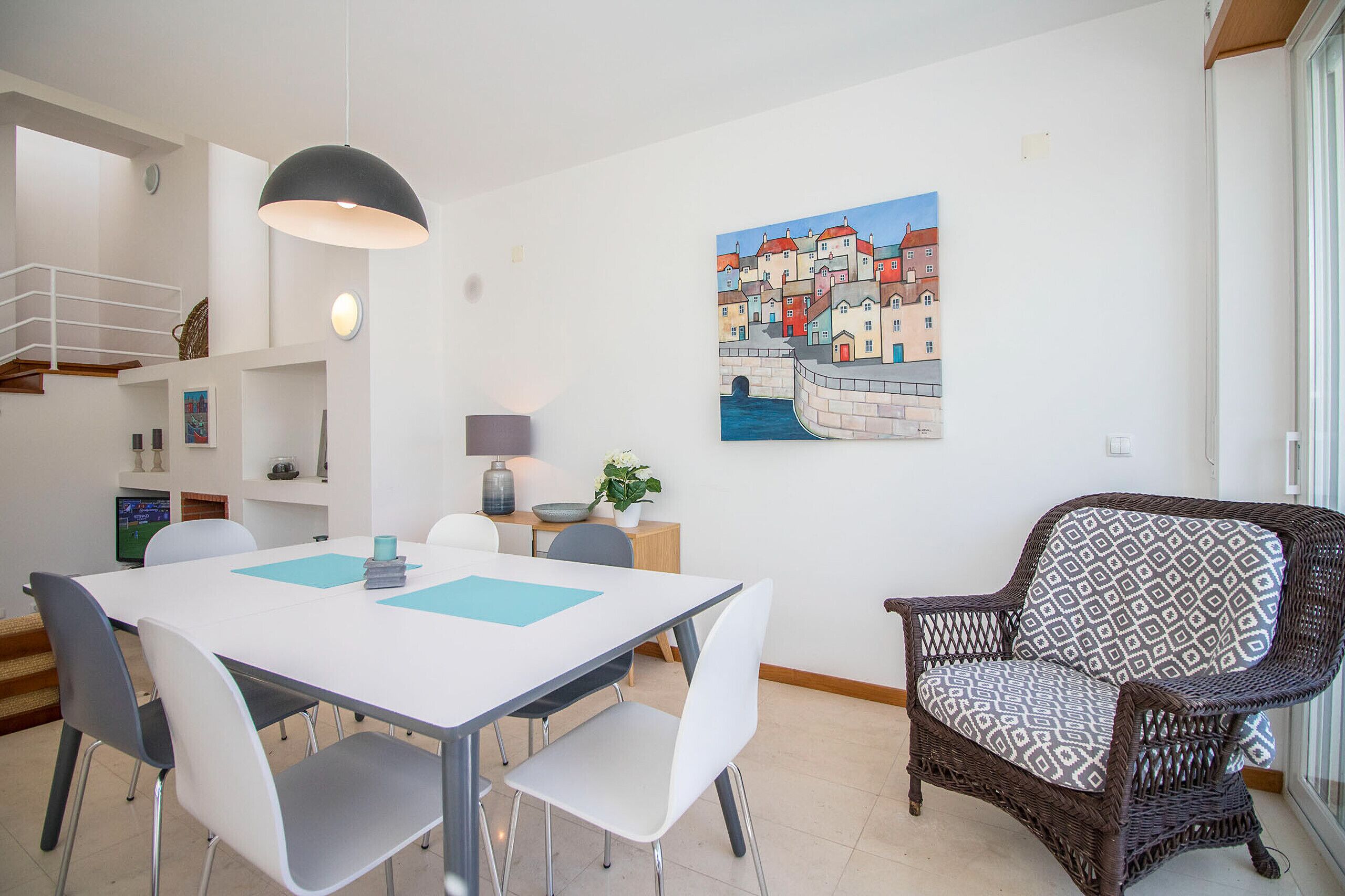 A dining area with a white table, grey chairs, a wicker chair, a painting of a town, and a hanging light fixture.