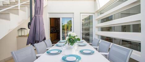 Een eetgedeelte met een witte tafel voor zes personen, grijze stoelen en een paarse parasol.