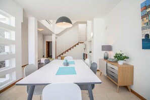 A modern dining area with a long table, chairs, a staircase, and a view of a building through large windows.