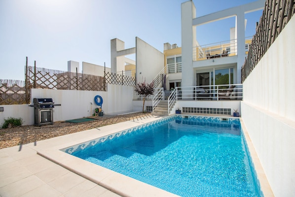 A swimming pool with a tiled edge, a barbecue grill, and a staircase leading to a balcony.