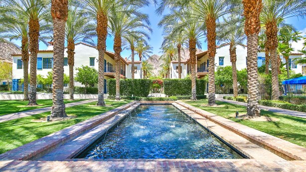 A rectangular pool with a brick border, surrounded by palm trees and a well-maintained lawn.