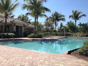 Une piscine avec une terrasse carrelée, des palmiers et, au fond, un bâtiment dont le toit est également recouvert de tuiles.