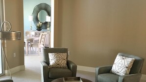 Ein Wohnzimmer mit zwei grauen Sesseln, einem runden Couchtisch und einem kleinen Beistelltisch. An der Wand hängt ein großer Spiegel, und im Hintergrund befindet sich ein Essbereich.