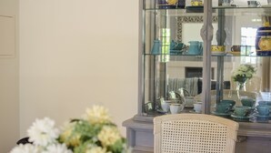 A room with a glass display cabinet containing various ceramic items, a wicker chair, and a vase with flowers.