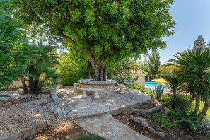 A stone bench area with a tree in the center, surrounded by a garden with various plants and a pool in the background. - Villa Caravela | 4 Bedrooms | Beautiful Garden | Almancil (Almancil)