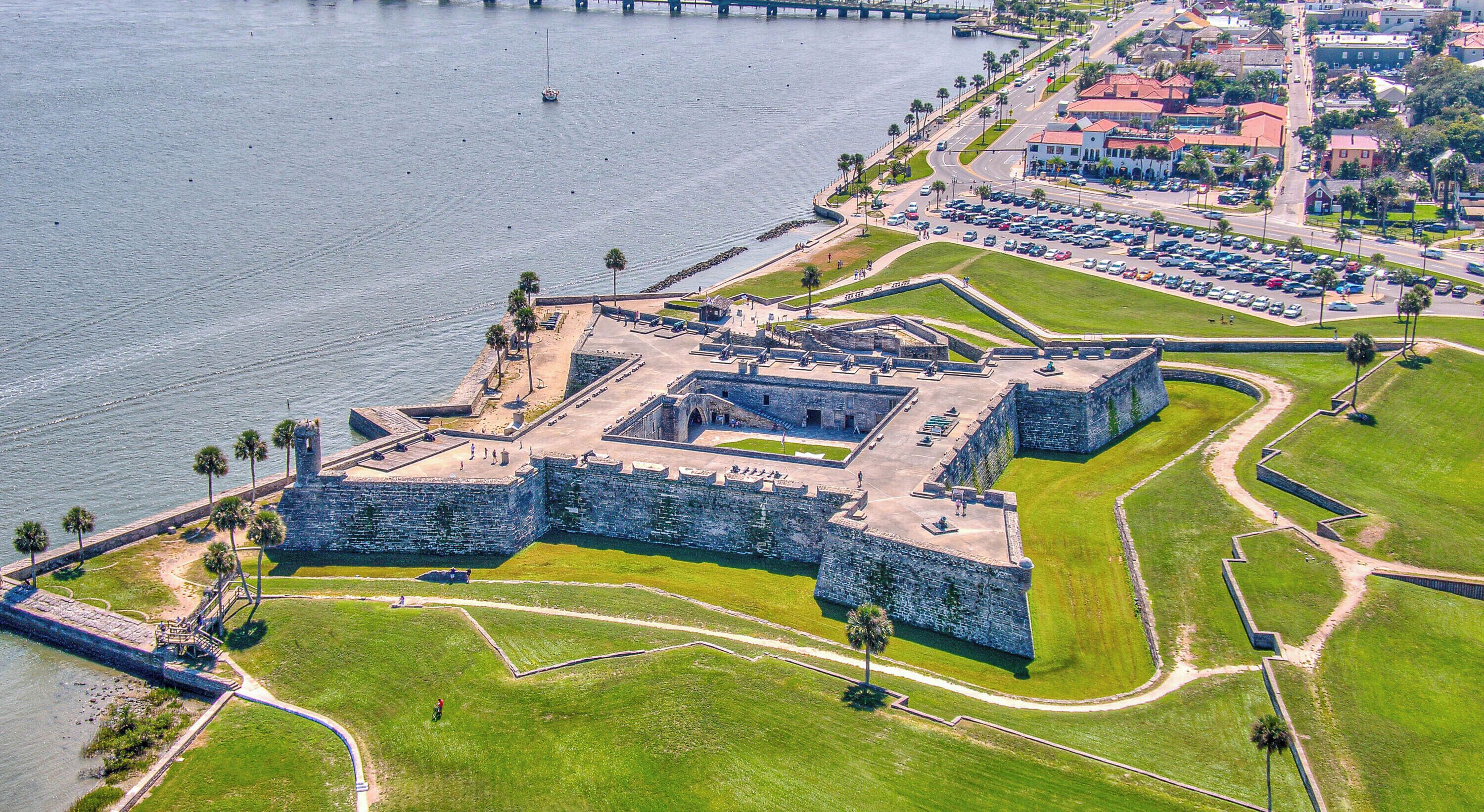 An aerial view of a historic fort with a large courtyard, surrounded by a well-maintained lawn and a body of water.