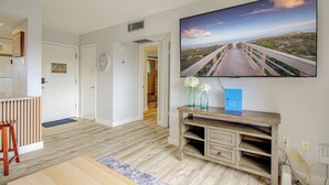 Un salon doté d’un grand téléviseur fixé au mur, d’un meuble de rangement en bois et offrant une vue sur l’extérieur par une fenêtre.