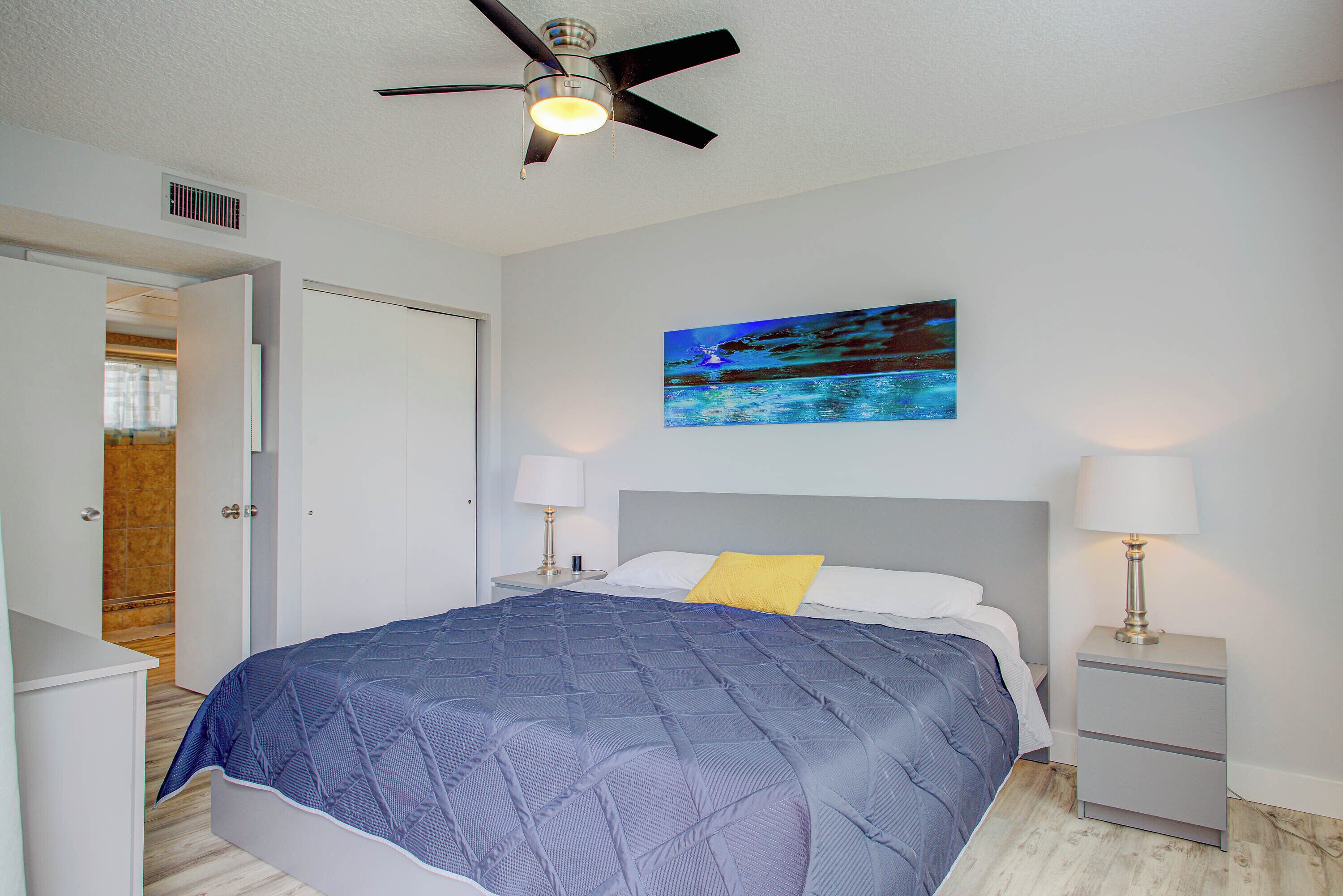 A bedroom with a bed, nightstands, a ceiling fan, a painting, and a door leading to another room.