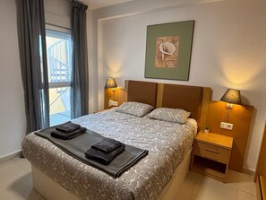 Un llit ben arreglat amb un edredó estampat i dues tovellones doblegades, una taula de nit amb una làmpada, un quadre amb marc a la paret i una finestra amb cortines.