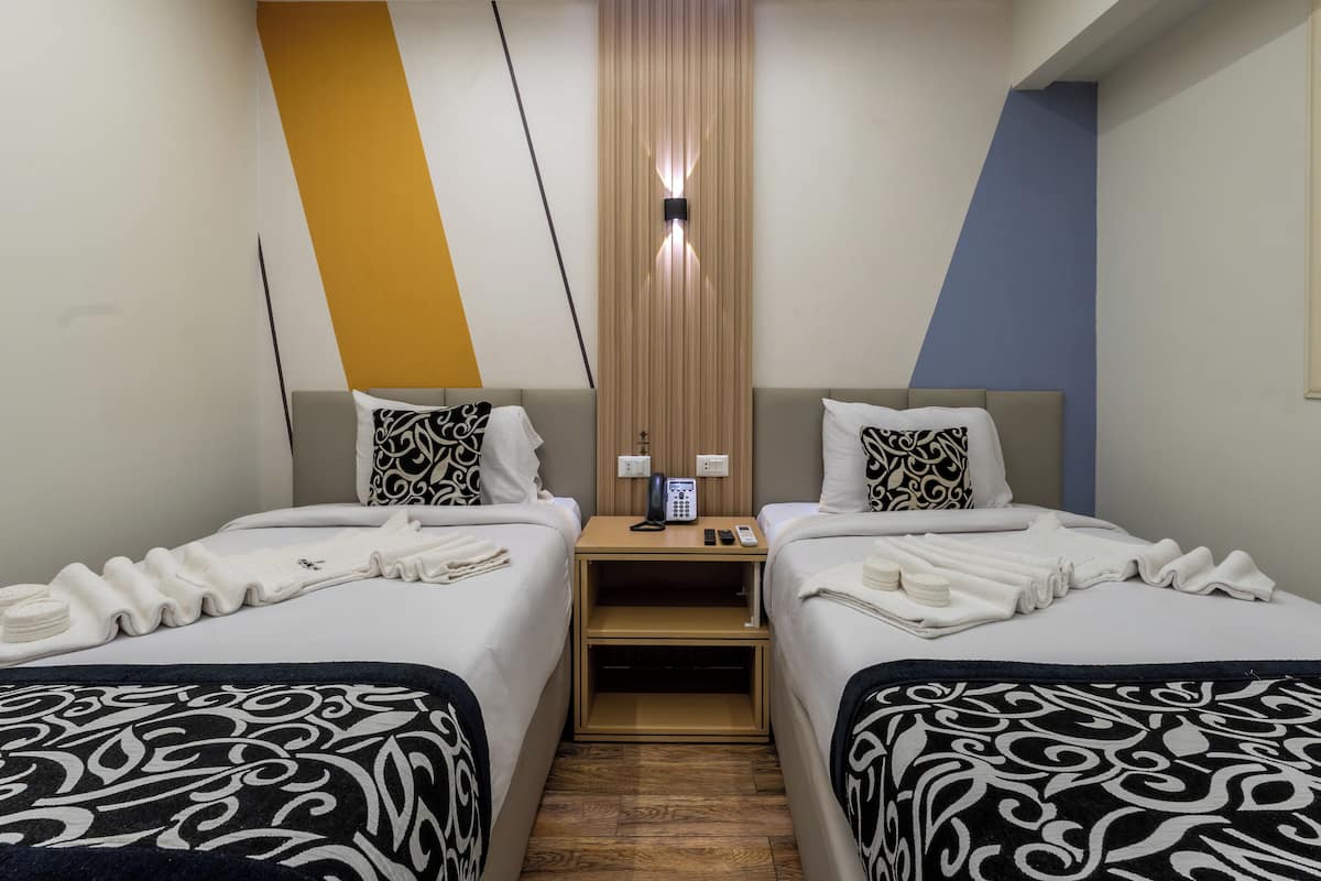 Habitación de hotel con dos camas, cada una con una colcha y almohadas estampadas, una mesita de noche de madera con un teléfono, y una pared con una lámpara vertical.