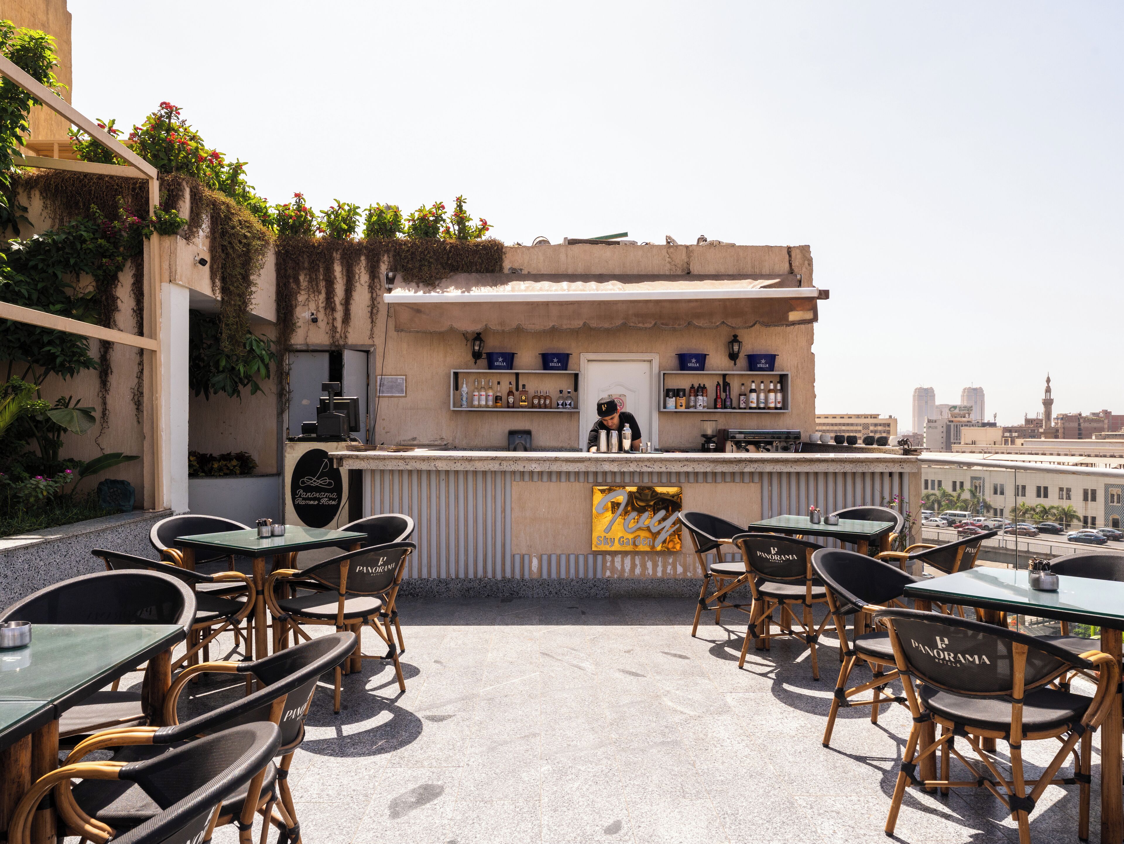 Um bar no terraço com mesas ao ar livre, um barman e o horizonte da cidade ao fundo.