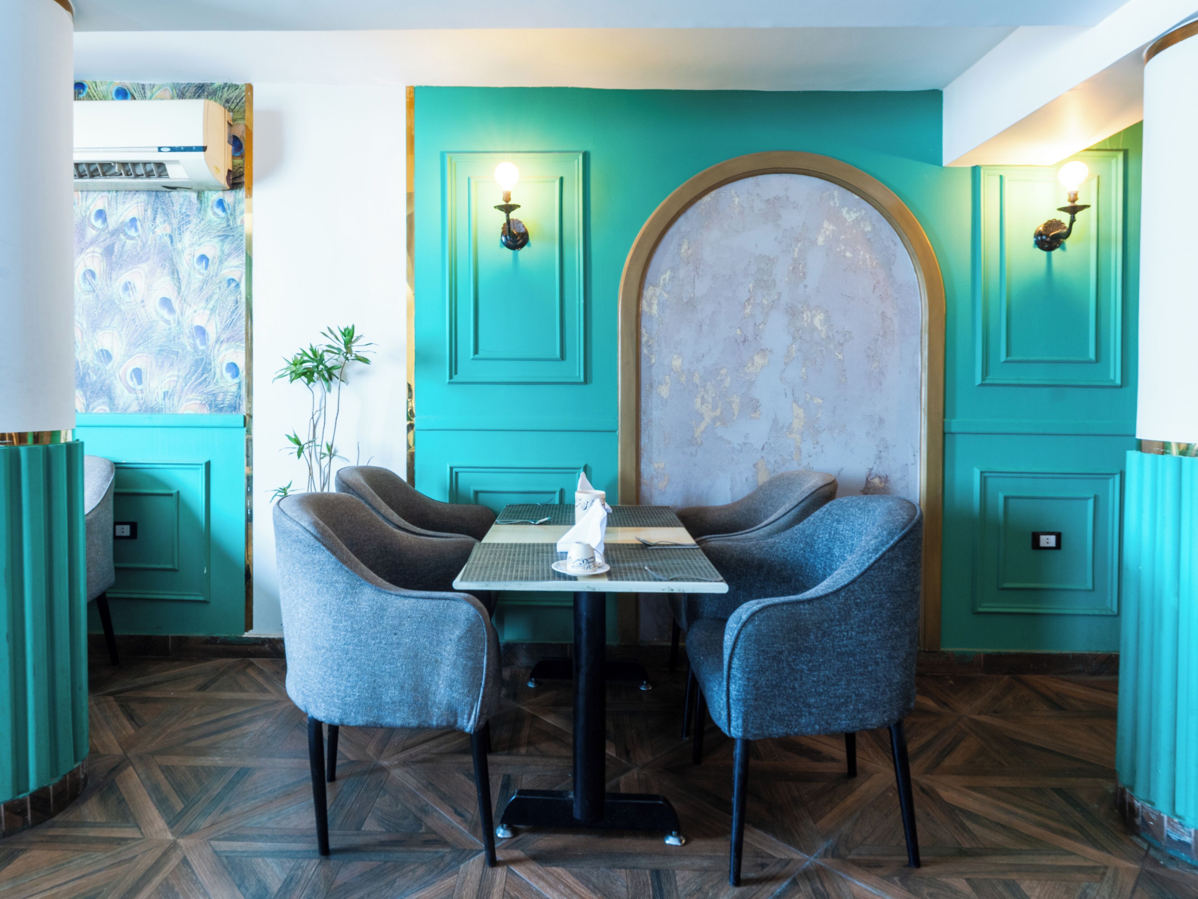 A dining area with teal walls, two grey upholstered chairs, a small table, and a decorative wall with peeling paint.