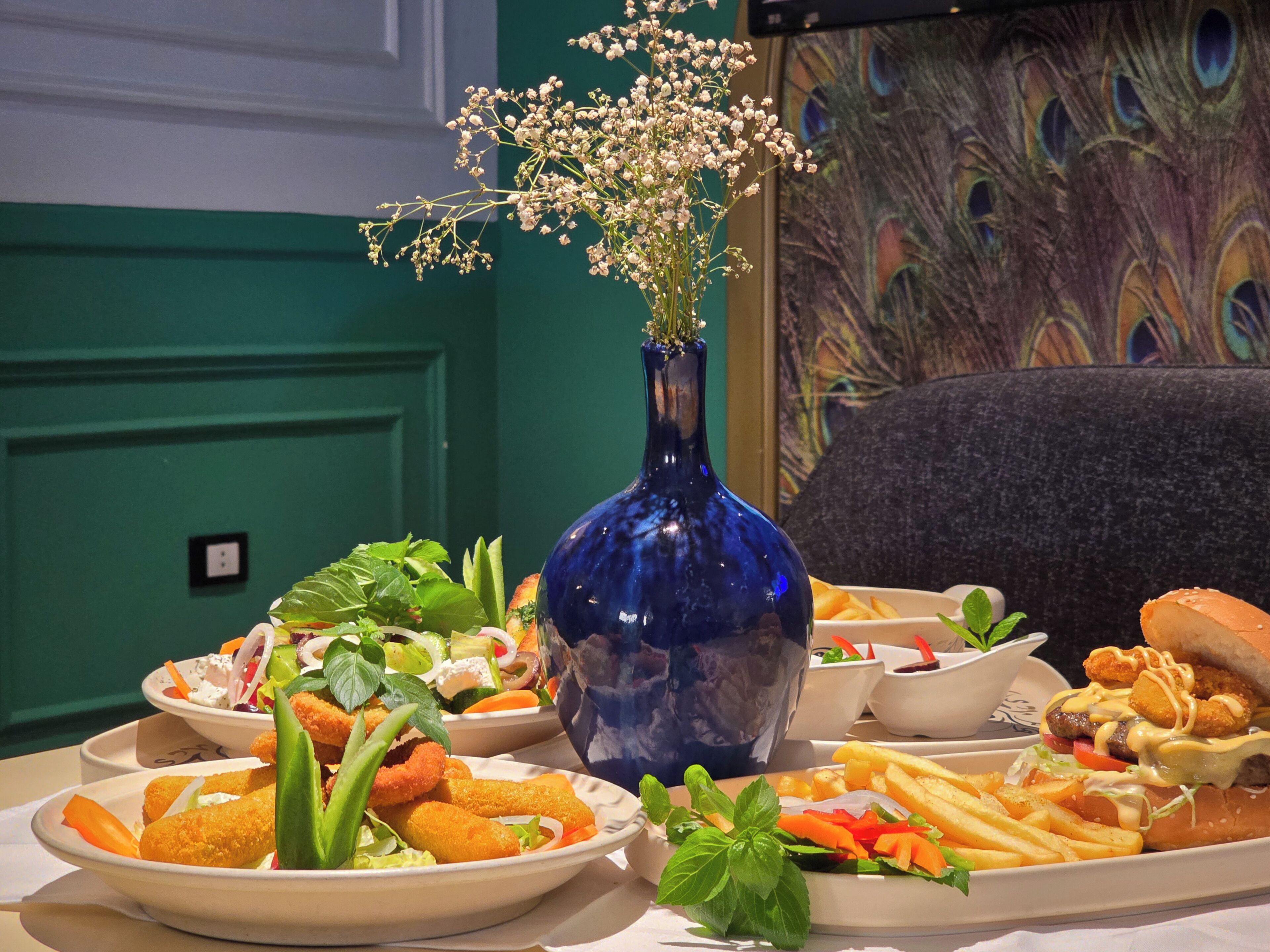 Une table garnie de différents plats, un vase bleu rempli de fleurs et un mur décoré sur le thème du paon.