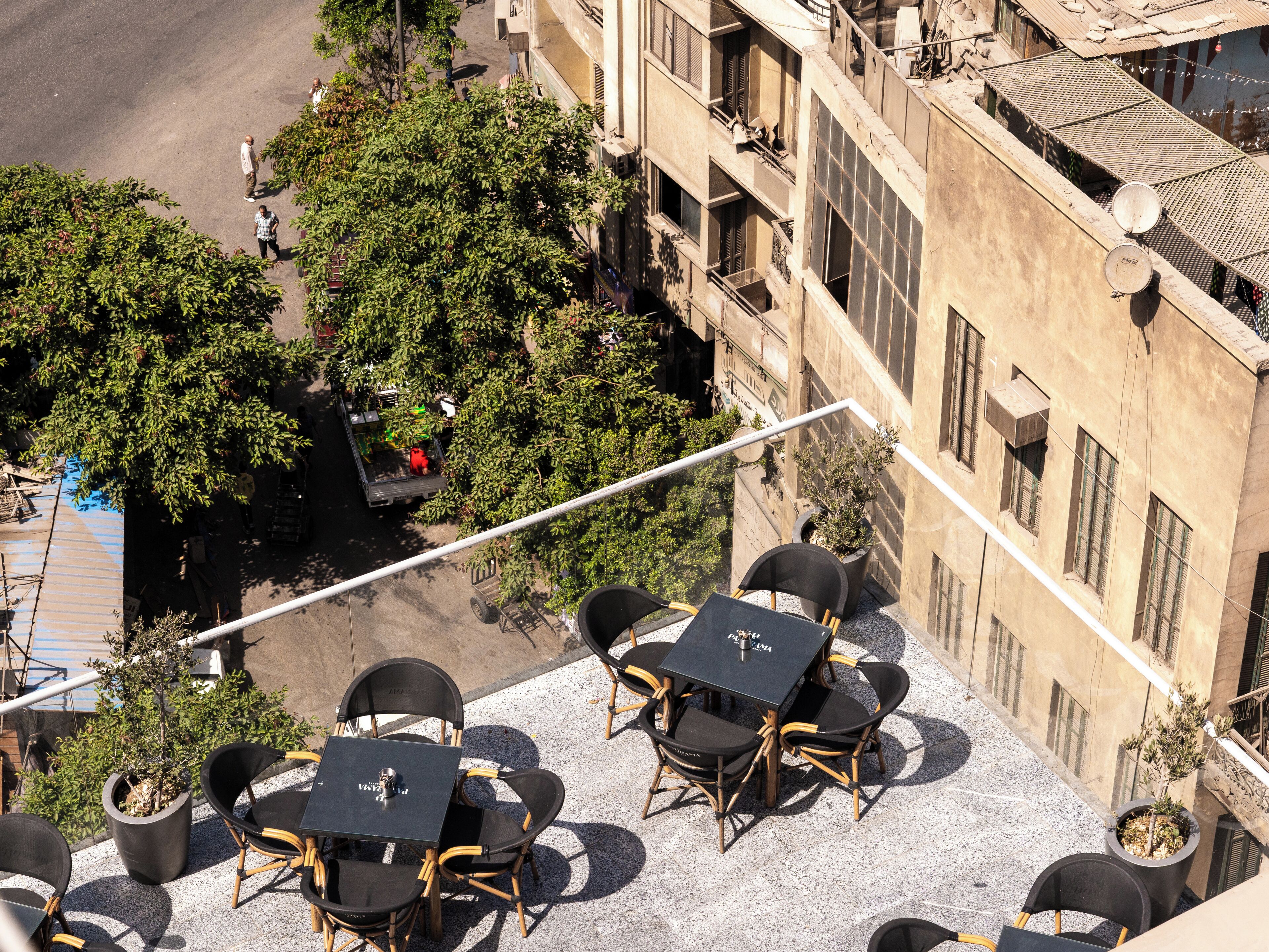 Une terrasse extérieure avec des tables et des chaises, entourée de verdure et bordée d’un bâtiment.