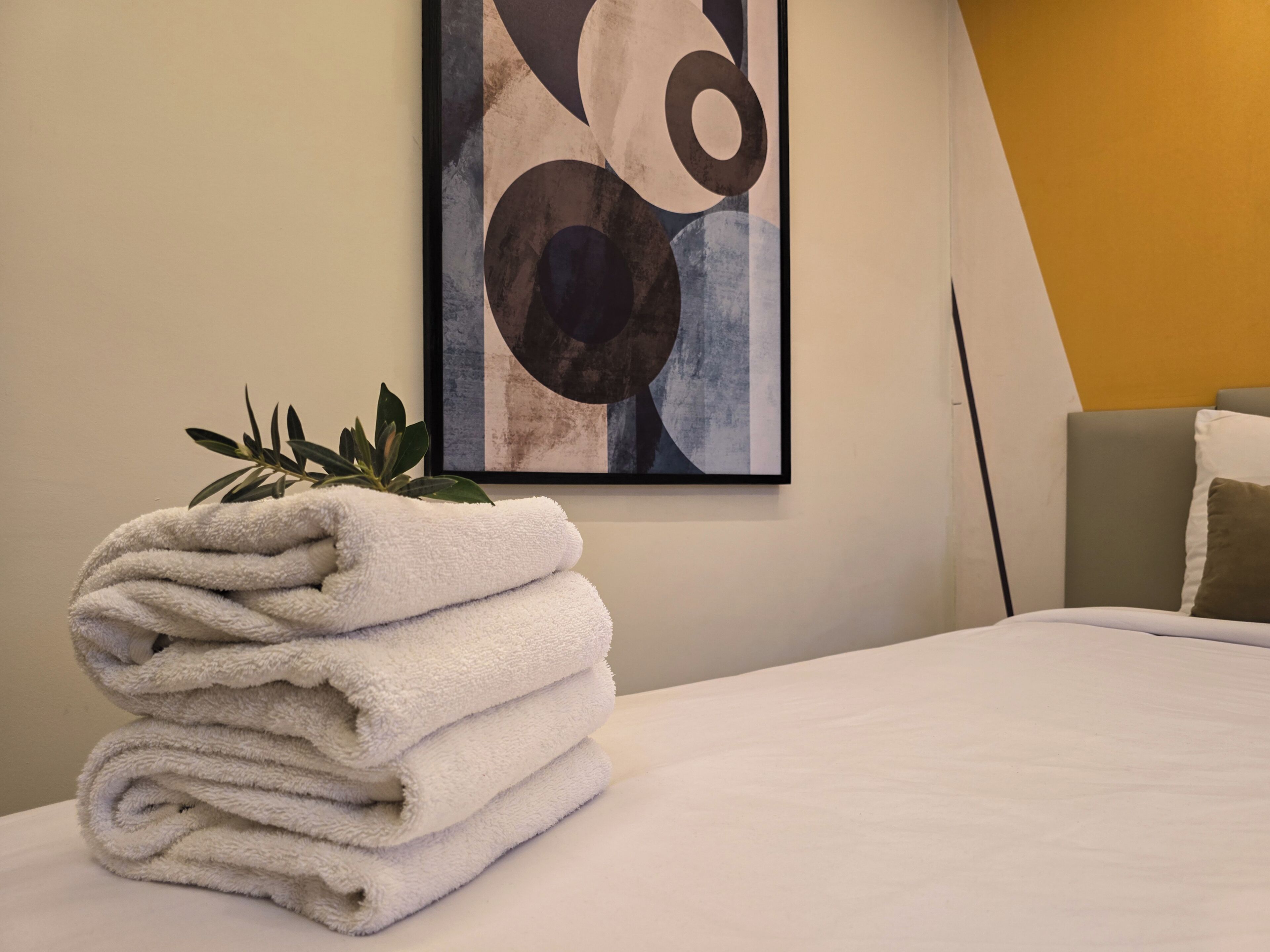 A neatly folded stack of white towels on a bed, a framed abstract painting on the wall, and a plant beside the towels.