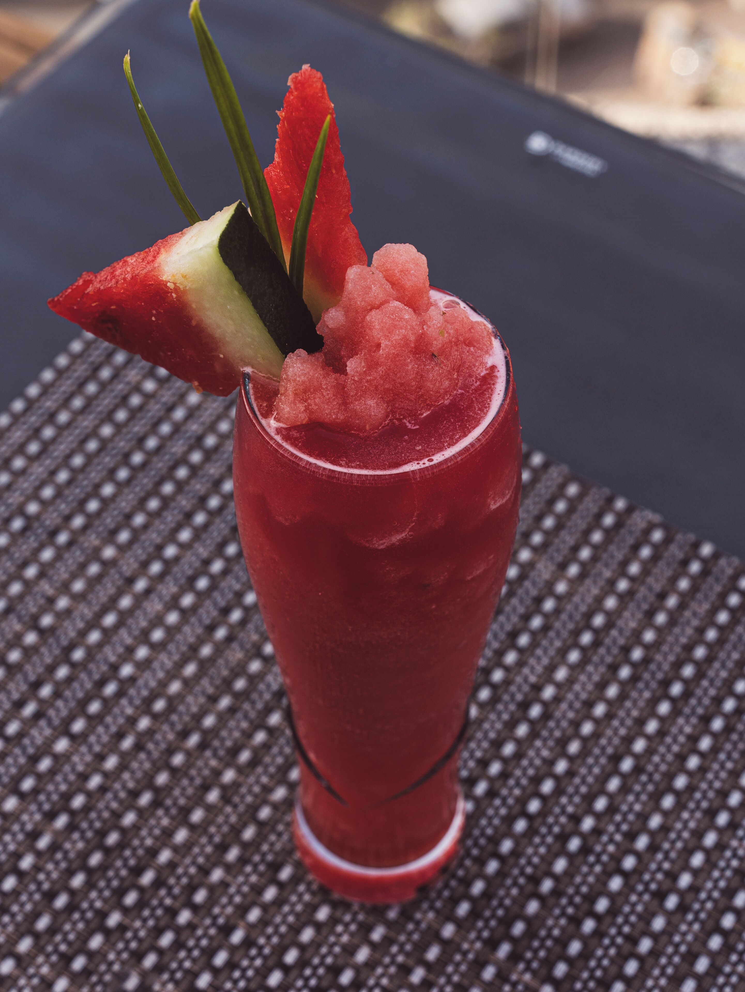 A tall glass with a red beverage, garnished with a slice of watermelon and a sprig of green herb, placed on a textured surface.