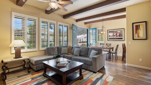A living room with a sectional sofa, a coffee table, and a colorful rug. There are large windows with blinds, a ceiling fan, and a dining area with a table and chairs. - Poolside! Stunning Villa near sparking pool & large spa, Garage, Pet Friendly! (L62-2) (La Quinta)