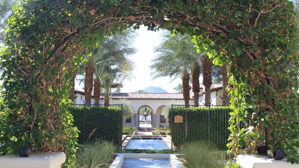 A pathway leading to a pool with a fountain, surrounded by lush greenery and palm trees, and a building with a tiled roof in the background.