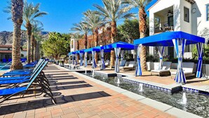 A poolside area with blue lounge chairs and umbrellas, palm trees, and a building with a balcony.
