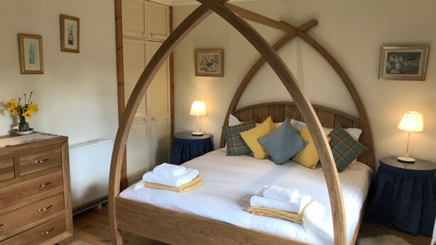 A wooden four-poster bed with a white mattress and multiple pillows, a wooden dresser with drawers, and two bedside tables with lamps.