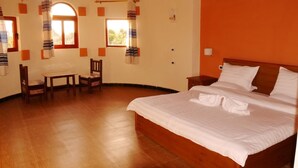 Habitación de hotel con piso de madera, una cama con sábanas blancas, una mesita de noche de madera, una silla, una pequeña mesa y dos ventanas con cortinas.
