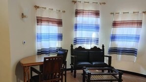 Una sala de estar con una mesa de comedor de madera, dos sillas, un sofá y una mesa baja. Hay tres ventanas con cortinas a rayas.