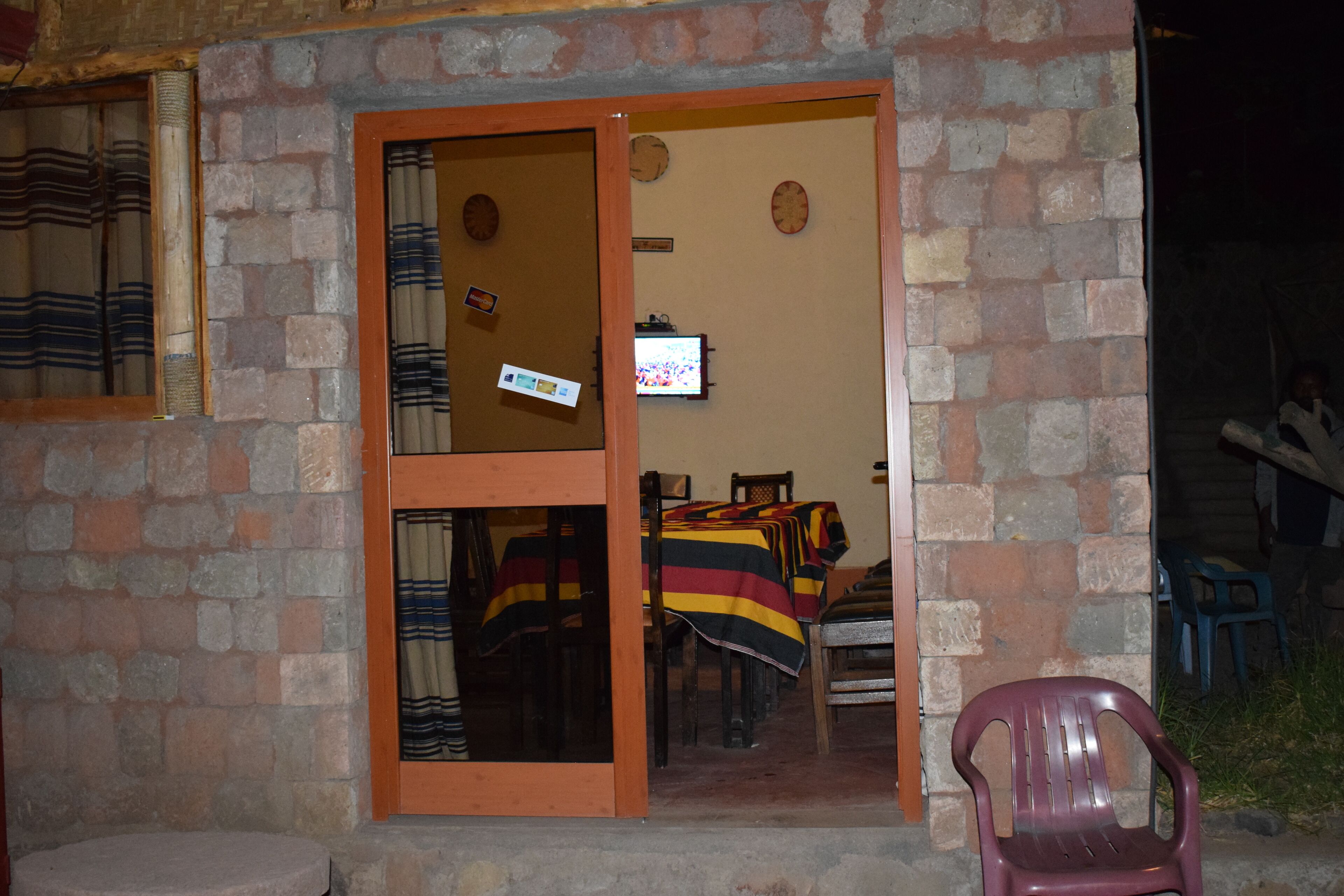 A brick building with a wooden door and windows. Inside, there are tables with striped tablecloths and chairs.
