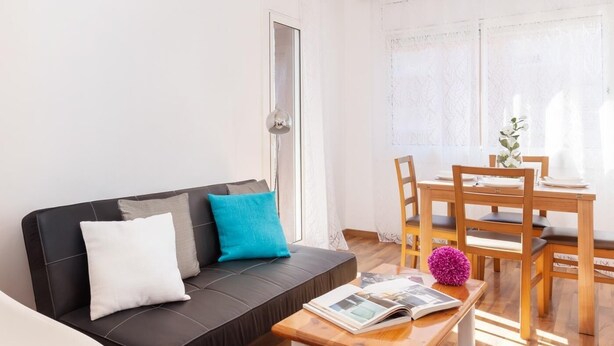 A modern living room with a black sofa, white and teal cushions, a wooden coffee table with a magazine, and a dining area with wooden chairs and a table. - Barcelona Fair 5min walking. TOP 300mb WiFi (L'Hospitalet de Llobregat)