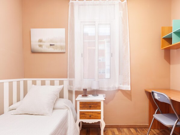 Un dormitorio con una cama, una mesita de noche con lámpara, una silla y una ventana con cortina.