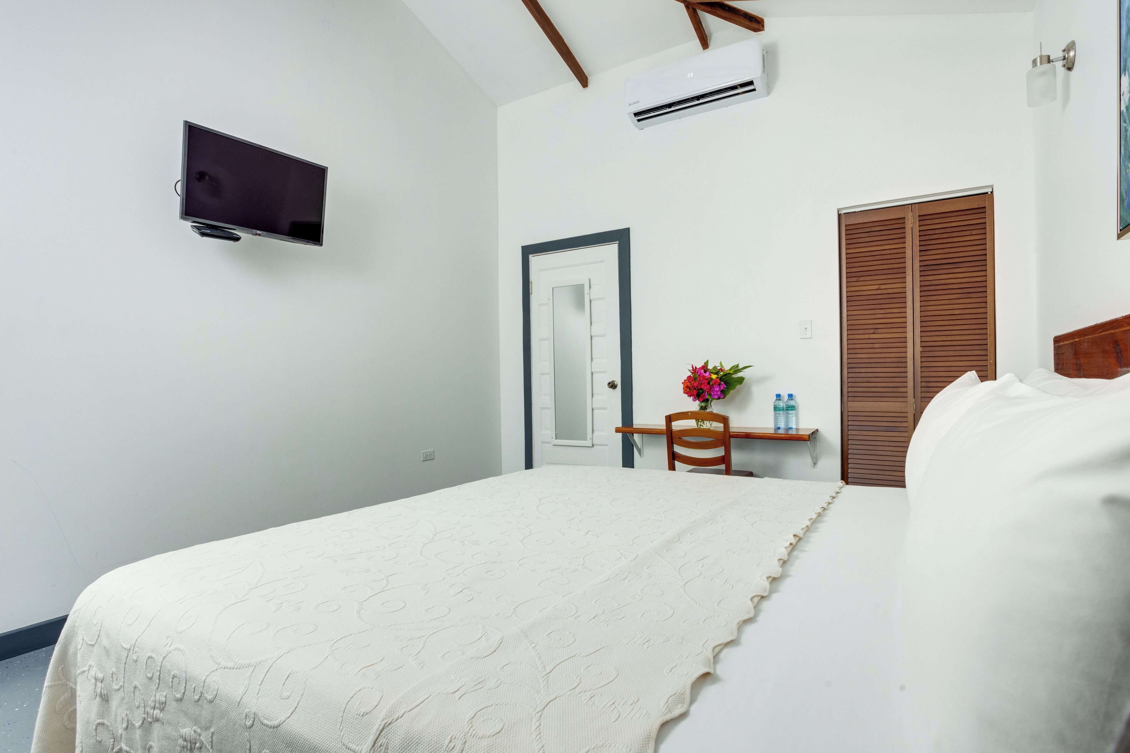 Une chambre à coucher avec un lit, un téléviseur fixé au mur, une porte en bois et une petite table sur laquelle est posé un vase de fleurs.