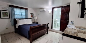 Habitación de hotel con cama, mesita de noche con lámpara, baño visible a través de una puerta de vidrio y suelo de baldosas.