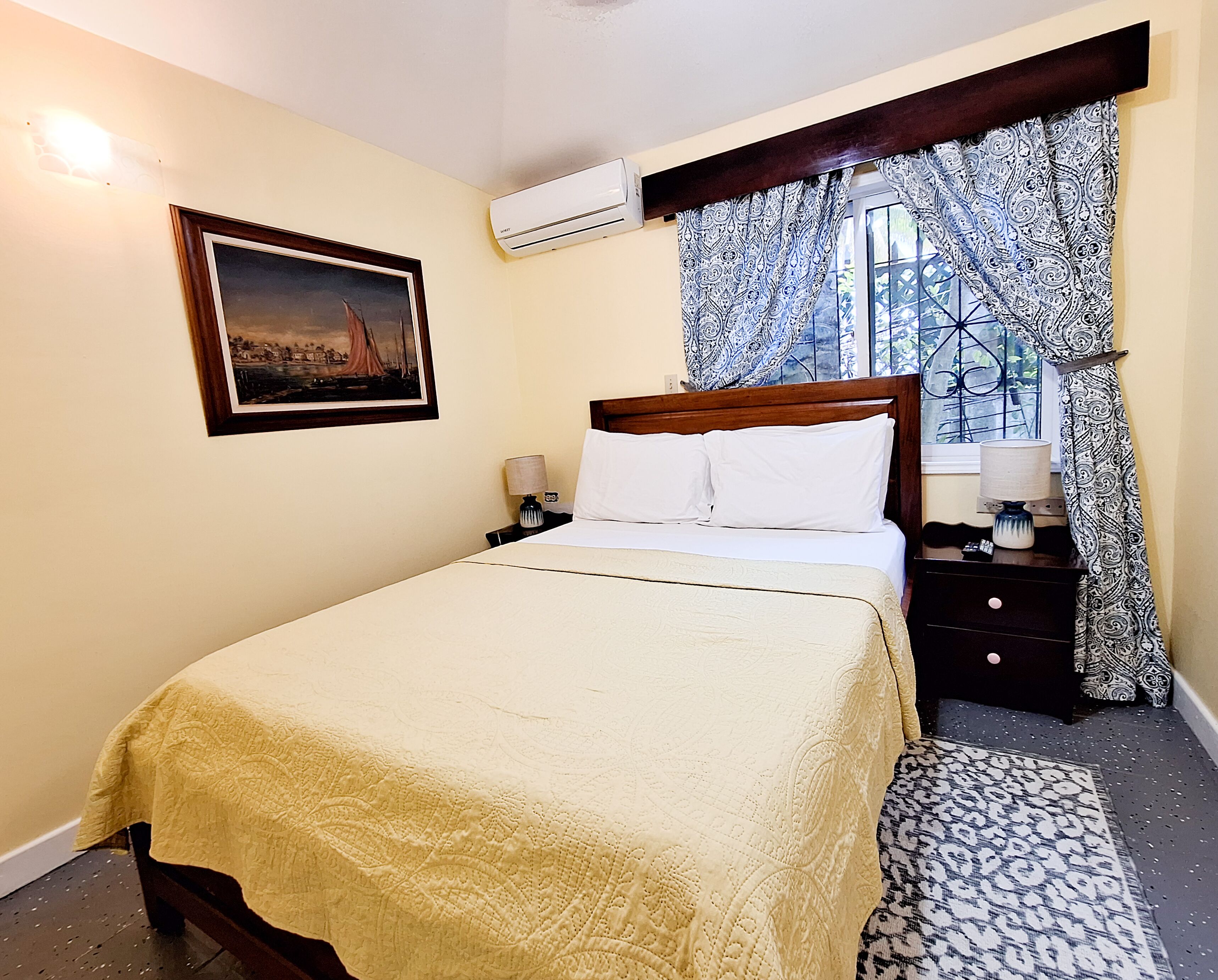 A neatly made bed with a white comforter and pillows, a bedside table with a lamp, a framed picture on the wall, and a window with patterned curtains.
